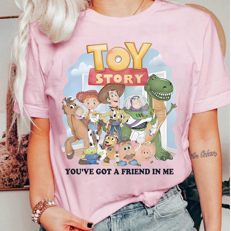 May include: Pink t-shirt featuring a graphic of characters from the animated film "Toy Story." The design includes Woody, Buzz Lightyear, Rex, and other characters, with the words "TOY STORY" and "YOU'VE GOT A FRIEND IN ME."