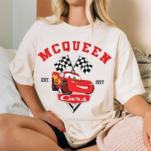 May include: Cream-colored t-shirt featuring a graphic of Lightning McQueen from the movie Cars. The design includes the text "MCQUEEN" in red, two checkered flags, and the word "Cars" in a white triangle.