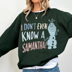 May include: Dark green sweatshirt with the phrase "I DON'T EVEN KNOW A SAMANTHA!" in pastel colors. A cartoon image of Olaf the snowman is on the right side of the sweatshirt. The sweatshirt has a white collar.