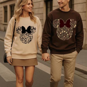 Puede incluir: Dos personas con sudaderas a juego. Una es beige, la otra marr&oacute;n. Cada sudadera presenta un dise&ntilde;o de cabeza de Mickey Mouse con estampado de leopardo y un lazo. La pareja se toma de la mano y camina por una calle.