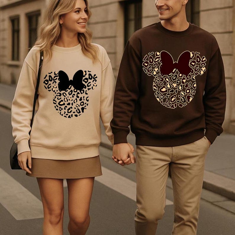 Puede incluir: Dos personas con sudaderas a juego. Una es beige, la otra marr&oacute;n. Cada sudadera presenta un dise&ntilde;o de cabeza de Mickey Mouse con estampado de leopardo y un lazo. La pareja se toma de la mano y camina por una calle.