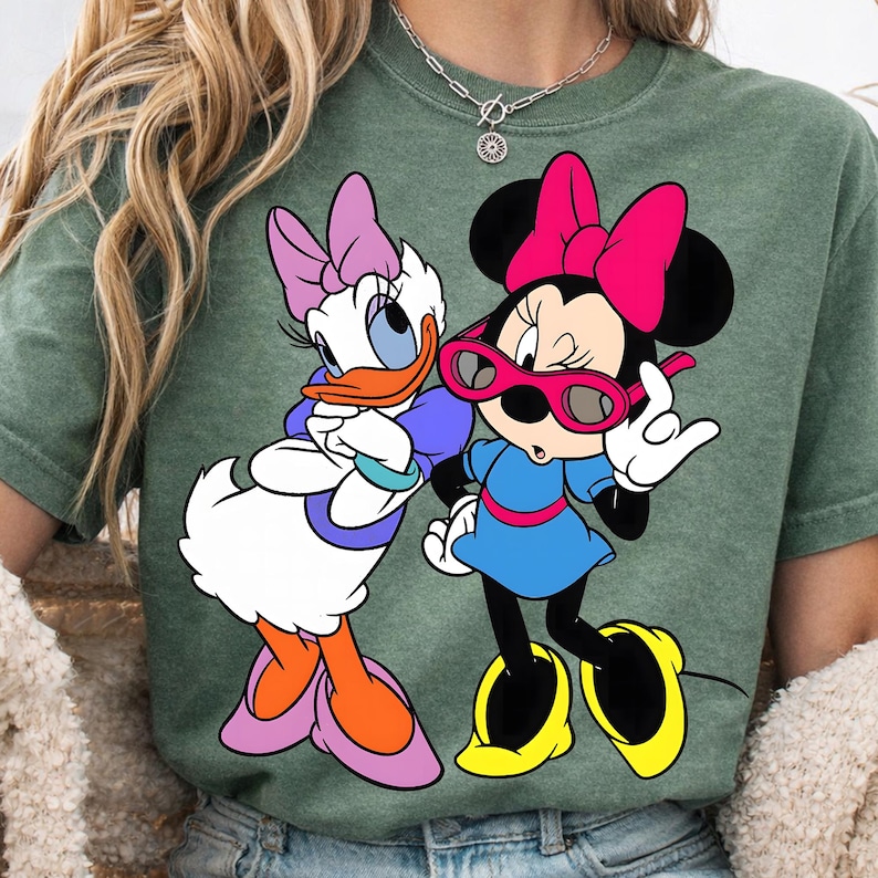 May include: Green t-shirt featuring Daisy Duck and Minnie Mouse cartoon characters. Daisy Duck is wearing a purple bow, and Minnie Mouse is wearing a pink bow and sunglasses. Both characters are in a colourful graphic design.