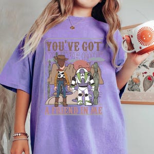 May include: A lilac-colored t-shirt featuring a graphic of two cartoon characters, a cowboy and a space ranger, with the text "YOU'VE GOT A FRIEND IN ME". The design includes desert scenery and a brown rectangular frame.