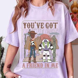 May include: A light purple t-shirt featuring a graphic of two cartoon characters, a cowboy and a space ranger, with the text "YOU'VE GOT A FRIEND IN ME". The design includes desert landscape elements.