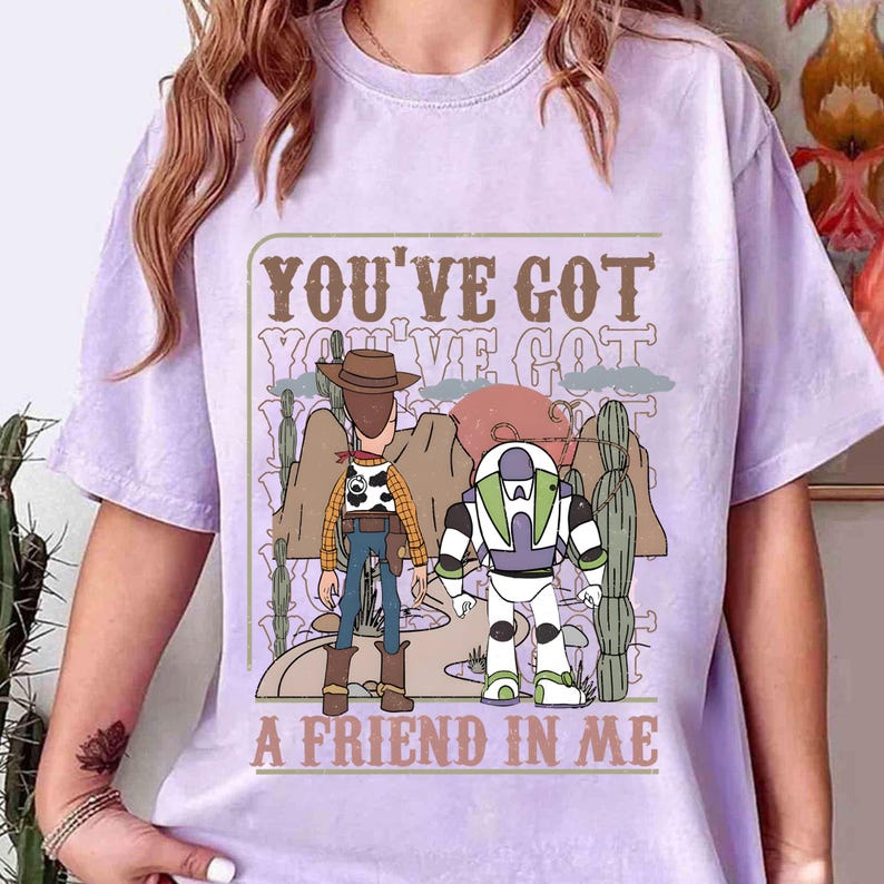 May include: A light purple t-shirt featuring a graphic of two cartoon characters, a cowboy and a space ranger, with the text "YOU'VE GOT A FRIEND IN ME". The design includes desert landscape elements.