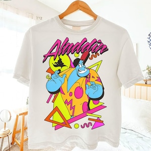 Op de afbeelding: Wit t-shirt met een kleurrijke afbeelding van de Geest uit Aladdin. Het ontwerp bevat het woord "Aladdin" in roze, met geometrische vormen en levendige kleuren. De Geest wordt afgebeeld met een blauwe huid en een rode hoed.