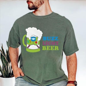 Op de afbeelding: Groen t-shirt met een cartoon bierpul ontwerp en de tekst "BUZZ LIGHT BEER". De bierpul is ontworpen om te lijken op het ruimtepak van Buzz Lightyear. Het shirt heeft een effen groene kleur.