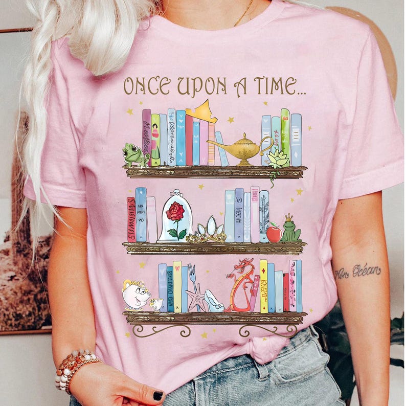 Może przedstawiać: R&oacute;żowa koszulka z napisem "ONCE UPON A TIME..." i wzorem p&oacute;łki na książki. P&oacute;łki są wypełnione kolorowymi książkami i ilustracjami z bajek, w tym żabą, r&oacute;żą i smokiem.