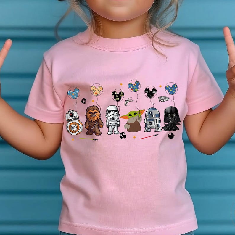 May include: Pink t-shirt featuring cartoon characters from Star Wars, including BB-8, Chewbacca, a Stormtrooper, Grogu, R2-D2, and Darth Vader. Each character is holding a balloon with a Mickey Mouse ear design. The shirt is a light pink color.