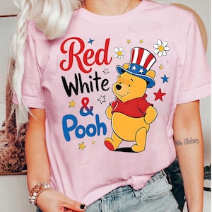Pode incluir: Uma camiseta rosa claro com o texto "Red White & Pooh" em vermelho, branco e azul. A camiseta apresenta um personagem de desenho animado de Winnie the Pooh usando um chapéu patriótico e uma camisa vermelha, com estrelas e flores.