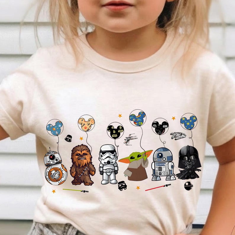 May include: Cream-colored t-shirt featuring a colorful graphic of Star Wars characters. The design includes BB-8, Chewbacca, a Stormtrooper, Grogu, R2-D2, and Darth Vader, each with a Mickey Mouse-themed balloon. Additional elements include stars and spaceships.