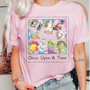 Op de afbeelding: Lichtroze T-shirt met een ingelijste afbeelding van stripfiguren. De afbeelding bevat een wasbeer, een tijger en andere personages. De tekst "Once Upon A Time" staat eronder gedrukt. Het shirt heeft korte mouwen.