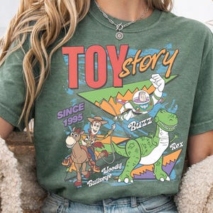May include: Green t-shirt featuring the Toy Story logo and characters. The design includes Woody, Buzz Lightyear, Rex the dinosaur, and Bullseye. The text "Since 1995" is also present. The shirt has a vintage, distressed look.