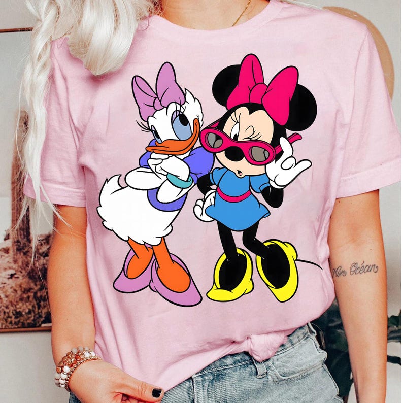 May include: Pale pink t-shirt with a cartoon graphic of Daisy Duck and Minnie Mouse. Daisy Duck is in a purple bow, blue top, and orange shoes. Minnie Mouse wears a pink bow, sunglasses, blue dress, and yellow shoes.
