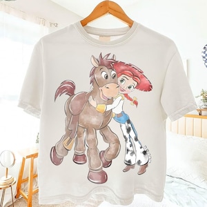 May include: Off-white t-shirt featuring a cartoon illustration of a character with red hair and a cowgirl outfit hugging a brown horse. The design is printed on a short-sleeved, crew neck tee shirt.