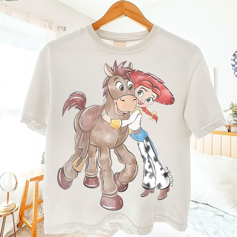 May include: Off-white t-shirt featuring a cartoon illustration of a character with red hair and a cowgirl outfit hugging a brown horse. The design is printed on a short-sleeved, crew neck tee shirt.
