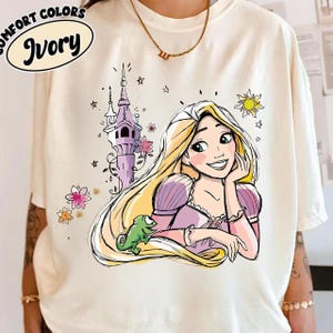 May include: Ivory-colored t-shirt featuring a cartoon illustration of a princess with long blonde hair, a green chameleon, and a castle. The shirt has the words "COMFORT COLORS" and "Ivory" printed on it.