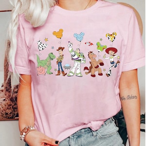 Puede incluir: Camiseta rosa claro con un gr&aacute;fico de dibujos animados de personajes animados. El dise&ntilde;o incluye un dinosaurio, un cerdo, un vaquero, un astronauta, un caballo y una vaquera, cada uno sosteniendo un globo.