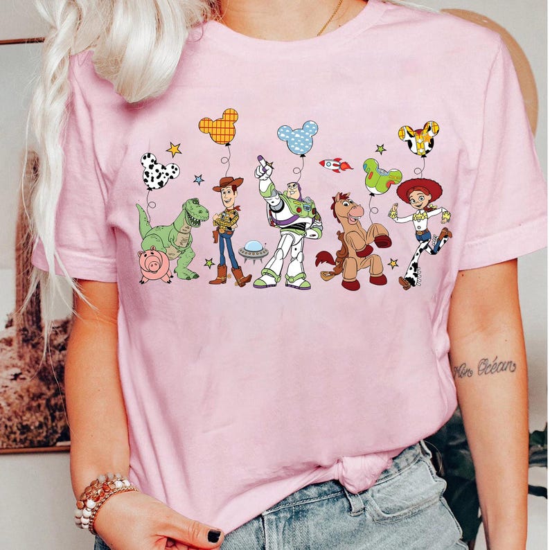 Puede incluir: Camiseta rosa claro con un gr&aacute;fico de dibujos animados de personajes animados. El dise&ntilde;o incluye un dinosaurio, un cerdo, un vaquero, un astronauta, un caballo y una vaquera, cada uno sosteniendo un globo.