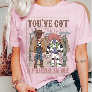 May include: Pink t-shirt featuring a graphic of Woody and Buzz Lightyear from Toy Story, with the text "YOU'VE GOT A FRIEND IN ME". The design includes a desert landscape with cacti and mountains.