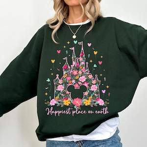 以下が含まれることがあります： 花柄の城のデザイン、ミッキーマウスの耳、そして「Happiest place on earth」の文字が特徴の濃い緑色のスウェットシャツ。 デザインには、ピンク、黄色、青の花、紙吹雪、星のアクセントが含まれています。 白い襟が見えます。