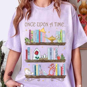 May include: A light purple t-shirt featuring a whimsical illustration of three bookshelves filled with storybook elements. The top shelf has a golden lamp and a frog. The middle shelf has a rose in a glass dome. The bottom shelf has a teacup and a dragon. The text "ONCE UPON A TIME..." is at the top.