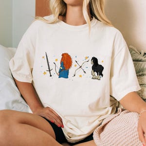 May include: Cream-colored t-shirt featuring a cartoon illustration of a sword, a character with red hair and a blue dress, a bow and arrow, and a black horse. The design includes yellow stars. The shirt is a casual, oversized fit.