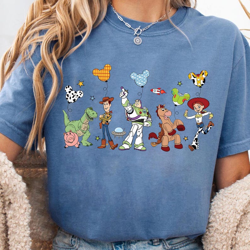 Puede incluir: Una camiseta azul con un gr&aacute;fico colorido de personajes de dibujos animados y globos. El dise&ntilde;o incluye un dinosaurio, un cerdo, un vaquero, un guardabosques espacial, un caballo y una vaquera. Los globos tienen varios patrones y formas.