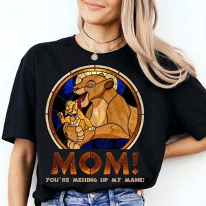 May include: Black t-shirt featuring a stained-glass style design of a lioness and cub. The design includes the text "MOM! YOU'RE MESSING UP MY MANE!" in a gold color. The shirt is a crew neck style.
