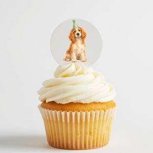 May include: A cupcake with white frosting and a round topper featuring a watercolor illustration of a Cocker Spaniel wearing a green party hat. The cupcake has a yellow cake base.