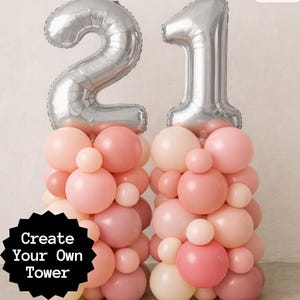 May include: Two balloon towers constructed from various shades of pink and peach latex balloons. Each tower is topped with a large silver foil balloon shaped as the numbers 2 and 1. A black starburst graphic includes the text Create Your Own Tower.