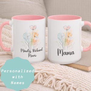 May include: Two white ceramic mugs with pink handles and interiors. Each mug features a floral design in pastel colors. One mug is inscribed with "Mindy, Richard, Mario", and the other with "Mama". The mugs are personalized with names.
