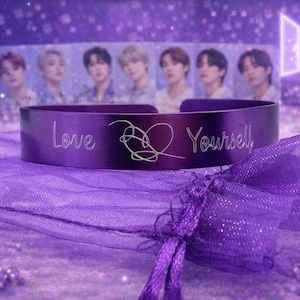 May include: A purple cuff bracelet with the words "Love Yourself" engraved on it, along with a heart-shaped design. The bracelet is displayed on a purple mesh bag, with a blurred background featuring images of people.