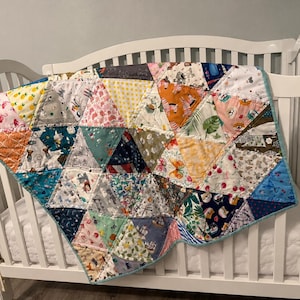 Puede incluir: Una colcha de patchwork que presenta una variedad de piezas de tela triangulares con diversos patrones, incluyendo flamencos, piñas, mariposas y varios animales en tonos rosa, amarillo, naranja, azul, verde y blanco con un borde azul claro.