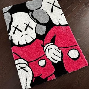 May include: A rectangular tufted rug featuring a graphic illustration of two cartoon characters with white skull-like heads, X-shaped eyes, and gloved hands. The characters are wearing bright pink shirts with white circular buttons against a black background.