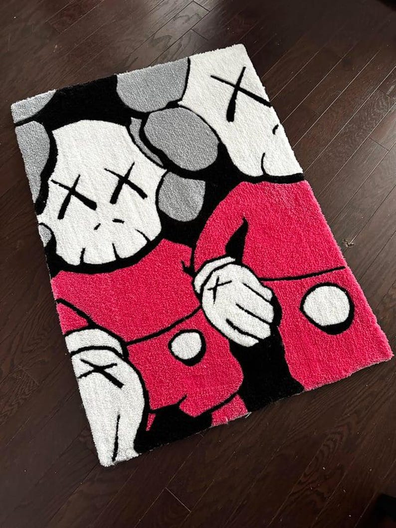 May include: A rectangular tufted rug featuring a graphic illustration of two cartoon characters with white skull-like heads, X-shaped eyes, and gloved hands. The characters are wearing bright pink shirts with white circular buttons against a black background.