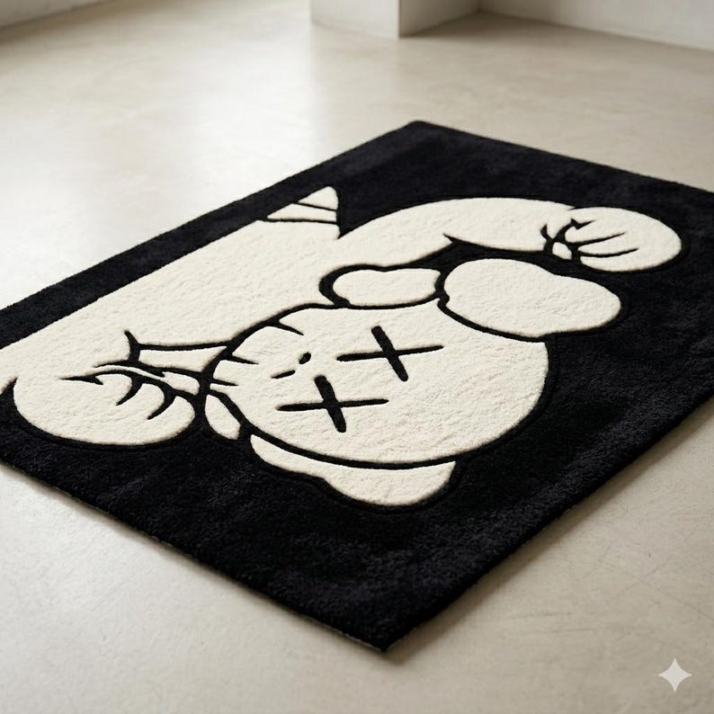 Puede incluir: Una alfombra rectangular con un dise&ntilde;o de alto contraste en blanco y negro. La alfombra muestra un personaje de dibujos animados grande y estilizado con extremidades redondeadas y ojos en forma de X, en blanco sobre fondo negro.