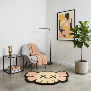 Puede incluir: Una alfombra de color crema y melocot&oacute;n con forma de cabeza de personaje estilizada, con contornos negros y ojos en forma de X. La alfombra est&aacute; sobre un suelo de hormig&oacute;n en una habitaci&oacute;n con un sill&oacute;n gris claro, una manta color melocot&oacute;n y una mesa auxiliar negra.