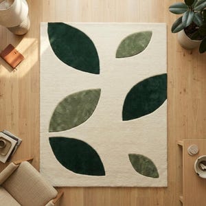 Puede incluir: Una alfombra rectangular con un fondo color crema y un patrón de grandes hojas verdes estilizadas. Las hojas varían en tonos de verde, creando un diseño texturizado y natural. La alfombra está colocada sobre un suelo de madera.