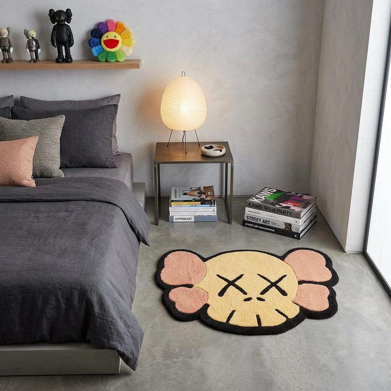 Puede incluir: Una alfombra con forma que presenta un rostro de personaje estilizado con un centro amarillo p&aacute;lido, acentos de color rosa claro en forma de orejas y contornos negros. El dise&ntilde;o incluye dos s&iacute;mbolos X negros para los ojos y una boca lineal.