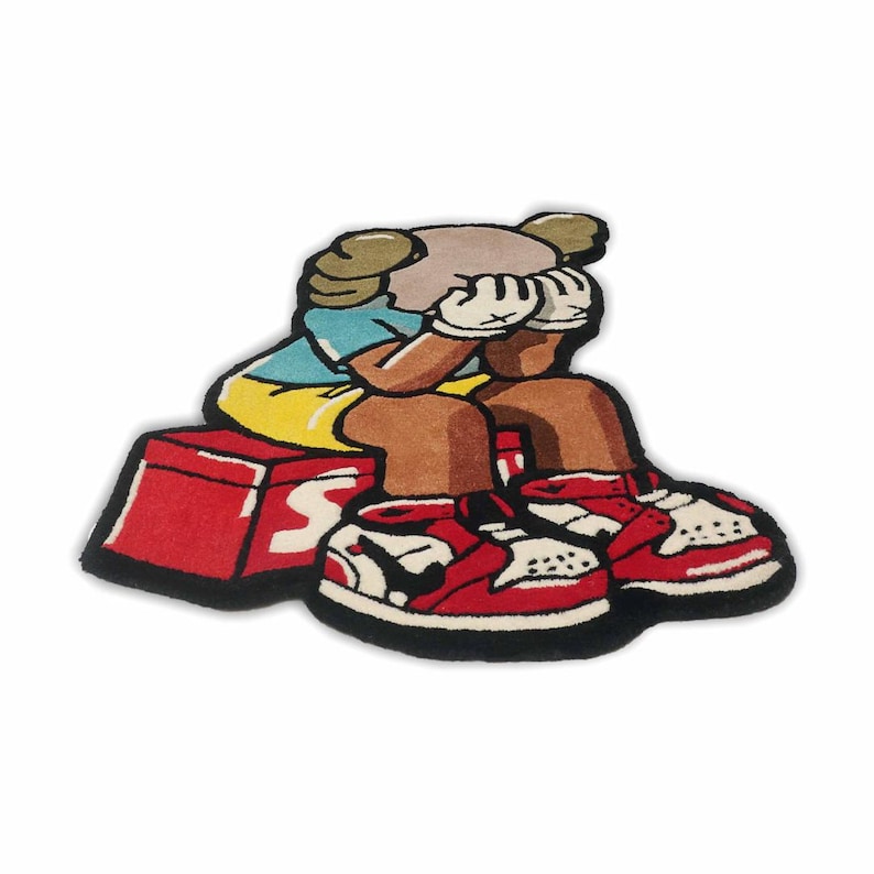 Puede incluir: Una alfombra de pelo cortado con forma de personaje de dibujos animados sentado sobre una caja roja. El personaje tiene tez bronceada, viste camiseta azul y pantalones cortos amarillos, y se cubre la cara con manos enguantadas blancas. Lleva zapatillas rojas y blancas.