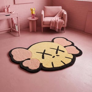 Puede incluir: Una alfombra de pelo insertado que presenta una cabeza de personaje estilizada con cara de color amarillo p&aacute;lido, orejas rosas y contornos negros. El dise&ntilde;o incluye dos ojos negros en forma de X y una peque&ntilde;a l&iacute;nea de boca negra en el suelo.