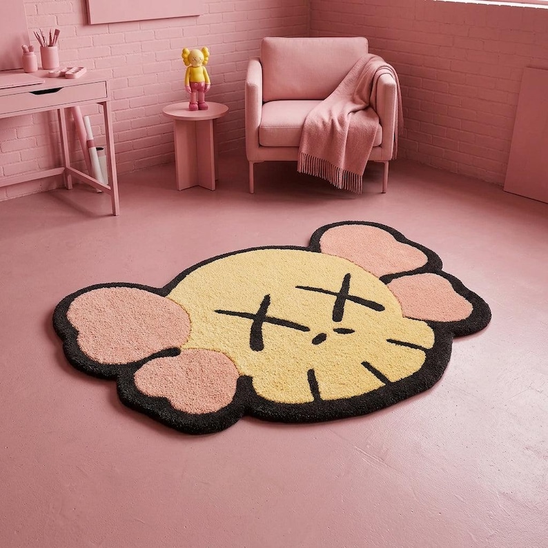 Puede incluir: Una alfombra de pelo insertado que presenta una cabeza de personaje estilizada con cara de color amarillo p&aacute;lido, orejas rosas y contornos negros. El dise&ntilde;o incluye dos ojos negros en forma de X y una peque&ntilde;a l&iacute;nea de boca negra en el suelo.