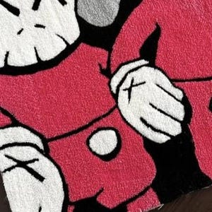 May include: A tufted rug featuring a graphic illustration of two cartoon-style figures with white heads, black X-shaped eyes, and white gloved hands. The figures are wearing red clothing with black accents, rendered in a textured, high-pile textile.