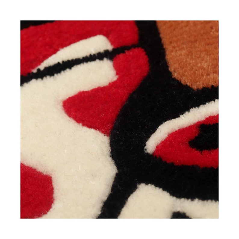 Puede incluir: Una superficie textil con mechones que presenta un patr&oacute;n gr&aacute;fico audaz con secciones de hilo negro grueso, rojo vibrante, blanco roto y bronceado. La textura es densa y afelpada, mostrando formas de colores con contornos definidos.