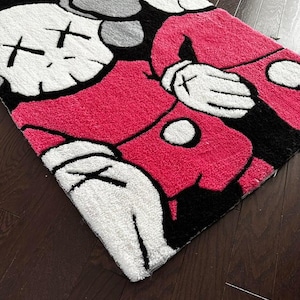 May include: A rectangular tufted rug featuring a stylized cartoon character with white gloved hands, a bright pink torso, and a white face with black X-shaped eyes. The design includes bold black outlines and a textured surface, showcasing a graphic pop art aesthetic with a high-contrast color palette of white, black, and vibrant pink.