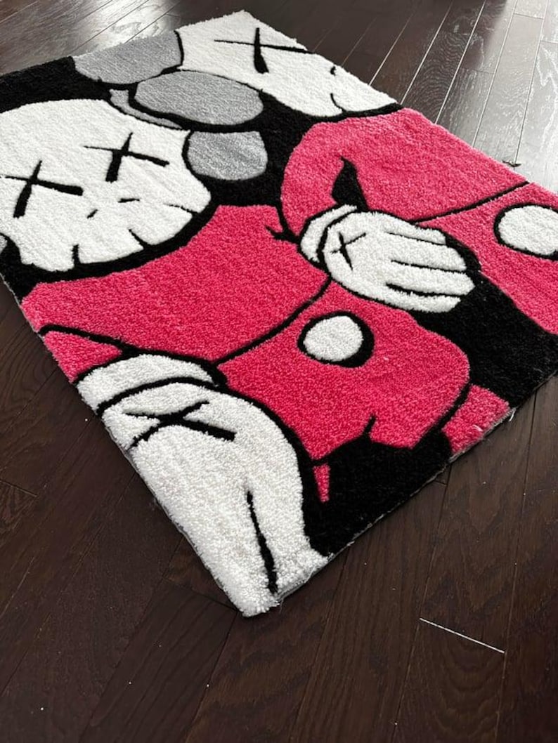 May include: A rectangular tufted rug featuring a stylized cartoon character with white gloved hands, a bright pink torso, and a white face with black X-shaped eyes. The design includes bold black outlines and a textured surface, showcasing a graphic pop art aesthetic with a high-contrast color palette of white, black, and vibrant pink.