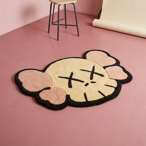 Puede incluir: Una alfombra de mechones con forma de cabeza de personaje estilizada, con un rostro color crema, ap&eacute;ndices rosados similares a orejas y contornos negros gruesos. Los rasgos faciales incluyen dos grandes ojos en forma de X y una l&iacute;nea negra simple para la boca, todo elaborado en un material texturizado y afelpado.