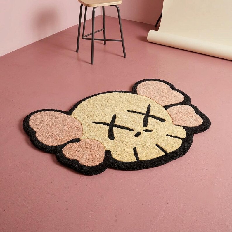 Puede incluir: Una alfombra de mechones con forma de cabeza de personaje estilizada, con un rostro color crema, ap&eacute;ndices rosados similares a orejas y contornos negros gruesos. Los rasgos faciales incluyen dos grandes ojos en forma de X y una l&iacute;nea negra simple para la boca, todo elaborado en un material texturizado y afelpado.