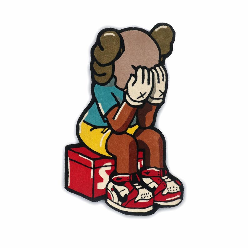 Puede incluir: Una alfombra de tufting con un personaje de dibujos animados de cabeza color canela y ojos en forma de X, que viste camiseta azul, pantalones cortos amarillos y guantes blancos. Est&aacute; sentado sobre una caja roja con texto blanco y zapatillas.